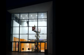 Zdjecie przedstawia dwóch pracowników, jeden stoi w podnośniku nożycowym, a drugi poza nim stojąc na szprosach myje elewacje szklaną łamiąc podstawowe zasady BHP.