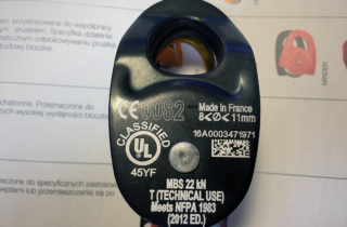Bloczek pojedynczy firmy Petzl koloru czarnego na którym widnieją białe oznaczenia mówiące o wytrzymałości, certyfikacji, przeznaczeniu oraz numer indywidualny produktu.