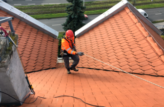 Pracownik pracujący na skośnym dachu wyposażony w środki ochrony indywidualnej - kask przemysłowy z czteropunktowym systemem mocowania, pełną uprząż z punktem wpięcia na klatce piersiowej i przyrząd autoasekuracyjny zainstalowany na linie półstatycznej.