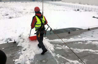 Pracownik ubrany w pełną uprząż, czerwony kask oraz kamizelkę odblaskową. Odśnieża dach budynku podpięty do systemu linowego chroniącego przed upadkiem.